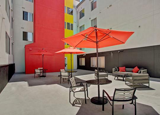 A modern outdoor courtyard here at Leilani on Greenwood showcasing a spacious concrete patio with café-style tables and chairs, bright red patio umbrellas providing shade, a comfortable outdoor sofa with accent pillows, and lounge seating, set against colorful building exteriors with bold red, yellow, and neutral tones that create a vibrant, urban atmosphere.