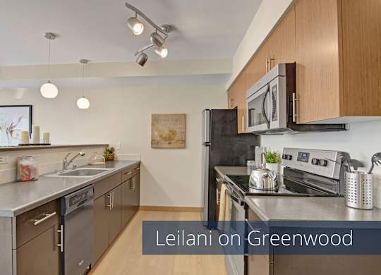 A modern galley-style kitchen here at Leilani on Greenwood featuring flat-panel wood cabinetry, smooth countertops, and a stainless steel sink with a dishwasher below. Dark appliances line the cooking area, while pendant and track lighting, light wood-style flooring, and a breakfast bar create a bright and functional space.