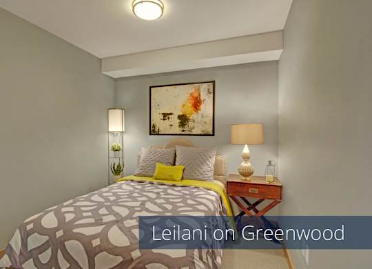 A cozy bedroom here at Leilani on Greenwood featuring soft carpeted flooring, neutral walls, and a bed with patterned bedding and accent pillows. A nightstand with a table lamp, framed artwork above the headboard, and overhead lighting create a warm, comfortable space designed for rest and relaxation.