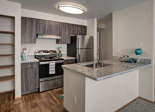 A modern kitchen here at Leilani on Greenwood featuring gray wood-style cabinetry, a light stone countertop, and a central island with an integrated sink and bar seating. Stainless steel appliances line the cooking area, while open shelving, wood-style flooring, and overhead lighting create a clean, contemporary space.
