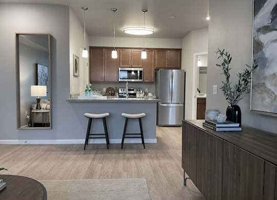 A modern open kitchen and living area here at LIVEasy Street featuring warm wood cabinetry, a stainless-steel refrigerator and range, a breakfast bar with two upholstered stools, pendant lighting, wood-style flooring, and a neutral living space with accent décor and soft natural light creating a welcoming apartment layout.