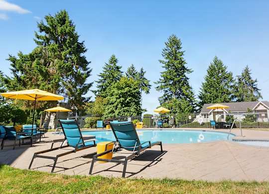 A sunny outdoor swimming pool here at Regency Woods showcasing a curved pool with a shallow entry, metal handrails, and surrounding concrete decking, complemented by teal lounge chairs, patio umbrellas, manicured lawns, tall evergreen trees, and a striped poolside cabana under a clear blue sky.