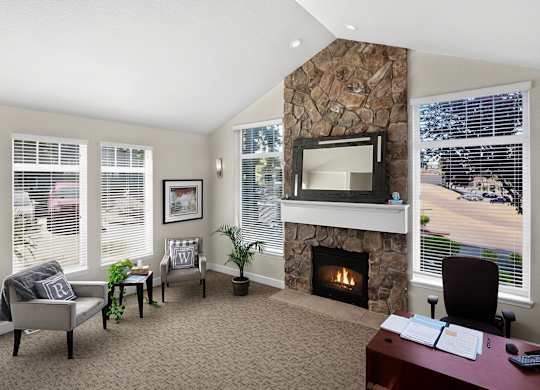 A bright business center here at Regency Woods featuring a stone fireplace with a mantel mirror, large windows with blinds allowing abundant natural light, neutral carpeting, a wooden desk with computer workstation, and cozy accent seating with side tables and plants, creating a comfortable and professional space for work or quiet meetings.