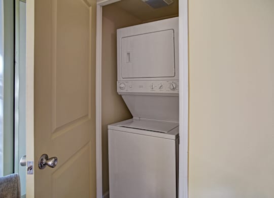 A compact in-home laundry closet here at Regency Woods featuring a stacked washer and dryer neatly tucked behind a paneled door, with neutral wall tones and a space-efficient layout that provides convenient, discreet laundry access within the apartment.