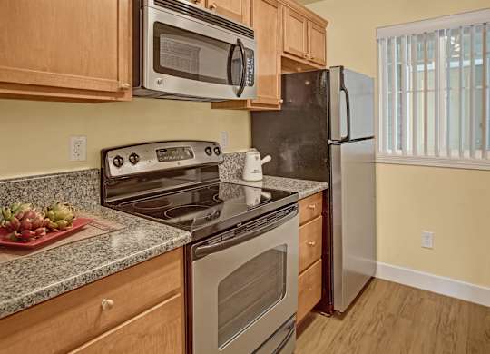 A warm kitchen here at Regency Woods featuring wood cabinetry, a full appliance suite, and speckled countertops, with a built-in microwave above the range, a refrigerator tucked beside a sunlit window with vertical blinds, and wood-style flooring that adds a clean, functional feel to the space.