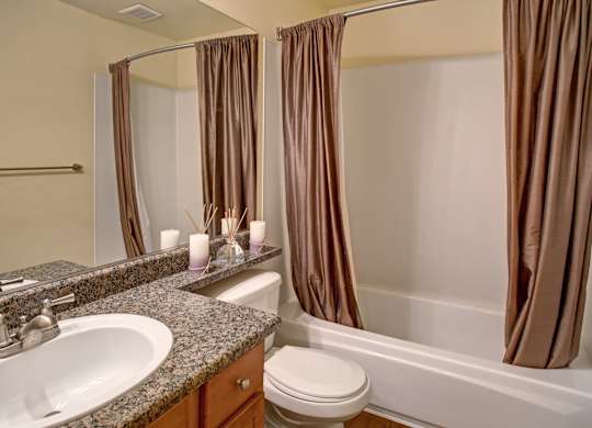 A clean bathroom here at Regency Woods featuring a wide granite-style vanity with an undermount sink, a large wall mirror, wood cabinetry, a toilet, and a tub with a shower curtain, complemented by neutral walls, simple décor accents, and durable flooring for a polished, functional space.