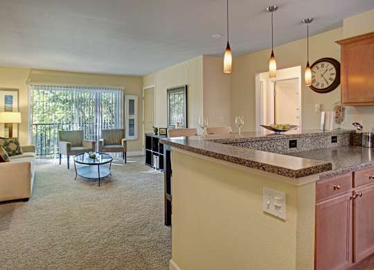 A welcoming open living and dining area here at Regency Woods showcasing a spacious carpeted layout with a comfortable seating area near a sliding glass door, a kitchen peninsula with speckled countertops and pendant lighting, wood cabinetry, and soft natural light that connects the kitchen and living spaces seamlessly.