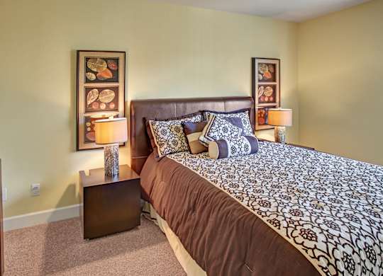 A comfortable bedroom here at Regency Woods featuring a spacious bed with patterned bedding, a padded headboard, matching nightstands with table lamps, framed wall art on either side of the bed, soft carpeted flooring, and light neutral walls that create a calm and inviting atmosphere.