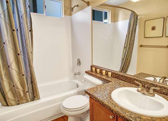 A clean bathroom here at Regency Woods featuring a full tub and shower with a curved curtain rod, granite-style vanity countertop with an integrated sink, wood-tone cabinetry, a large wall mirror, towel bar, and soft overhead lighting creating a bright and functional space.