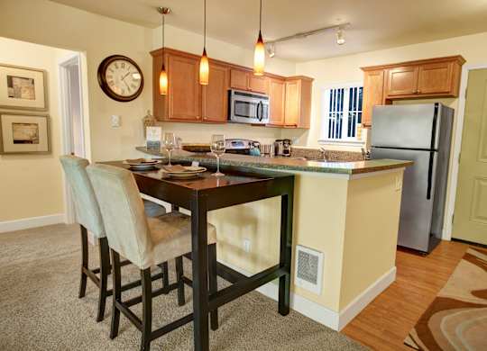 A welcoming kitchen and dining area here at Regency Woods featuring warm wood cabinetry, a full appliance suite, a breakfast bar with seating, a tall dining table with upholstered chairs, pendant lighting, wood-style flooring in the kitchen, soft carpet in the dining space, and a nearby entry door that adds everyday convenience.