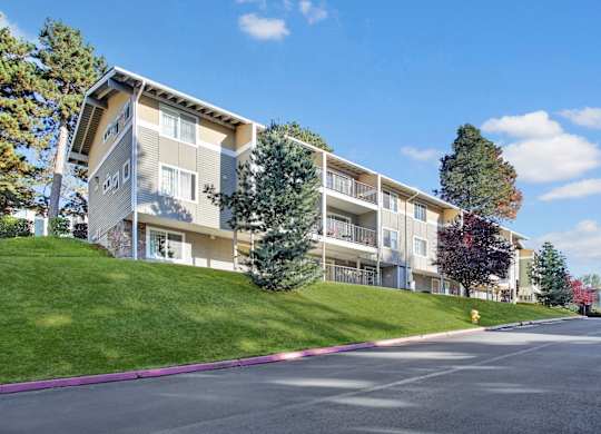 A hillside apartment building here at Regency Woods featuring multi-level residences with private balconies, neutral-toned siding, and large windows, set above a gently sloped green lawn with mature trees, seasonal foliage, and a paved driveway below under a clear blue sky with soft clouds.