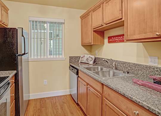 A cozy kitchen here at Regency Woods showcasing warm wood cabinetry, speckled countertops, and a double-basin sink beneath upper cabinets, with a dishwasher, refrigerator, and range arranged along the walls, wood-style flooring underfoot, and a window with vertical blinds bringing in soft natural light.
