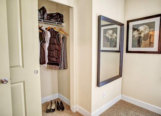 A compact entry closet area here at Regency Woods featuring an open hanging rod with shelving above, neatly arranged coats and jackets, carpeted flooring, and a recessed nook beside a full-length wall mirror and framed artwork, offering convenient storage and a clean, organized transition space near the entry.