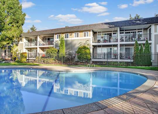A peaceful outdoor swimming pool here at Regency Woods showcasing a curved pool with calm blue water, a surrounding concrete deck, and a black metal fence, set in front of multi-story apartment buildings with private balconies, mature trees, landscaped greenery, and a bright blue sky creating a relaxed community atmosphere.