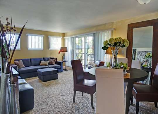 A spacious living and dining area here at Regency Woods featuring plush carpet flooring, a sectional sofa with an ottoman, a round dining table with mixed upholstered chairs, large windows with vertical blinds, and a sliding glass door leading to a private balcony, creating a warm, open layout filled with natural light.