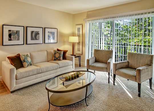 A bright living room here at Regency Woods featuring a neutral sofa with patterned accent pillows, two upholstered armchairs, and a glass coffee table set on plush carpeting, with framed wall art, a floor lamp, and large sliding glass doors with vertical blinds that open to a private balcony and bring in natural light.