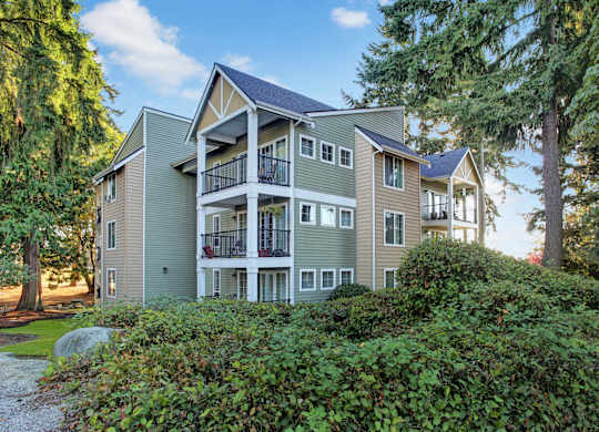 A landscaped exterior apartment building here at Regency Woods featuring three stories of residences with private balconies, neutral-toned siding, white trim accents, and black metal railings, set among manicured lawns, shrubs, and mature evergreen trees under a bright blue sky.