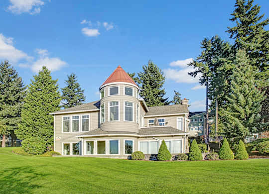 A distinctive clubhouse building here at Regency Woods featuring a rounded tower with a copper-toned roof, expansive windows, and light neutral siding, set against manicured green lawns, trimmed shrubs, and tall evergreen trees under a bright blue sky with scattered clouds.