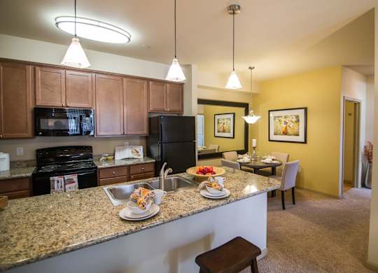 A welcoming kitchen here at Riverside 9 with a large granite breakfast bar, warm wood cabinets, black appliances, pendant lighting, and an open layout connecting to the dining area.