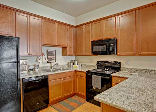 A bright kitchen here at Riverside 9 with warm wood cabinets, granite countertops, black appliances, and a functional L-shaped layout with ample counter space.