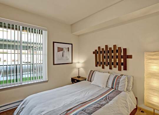 A cozy bedroom here at Riverside 9 with a comfortable bed, warm wood headboard accent, bedside table with lamp, and a large window with vertical blinds bringing in natural light.