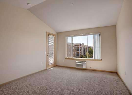 A spacious, empty living room here at The Vintage with soft beige carpet, light neutral walls, and a large window bringing in natural light. A glass door with blinds leads outside, and a wall-mounted air conditioner sits below the window.
