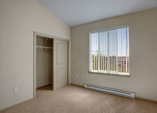 A bright bedroom here at The Vintage with soft beige carpet, light neutral walls, and a window with vertical blinds bringing in natural light. A spacious closet with sliding doors and built-in shelving provides convenient storage.
