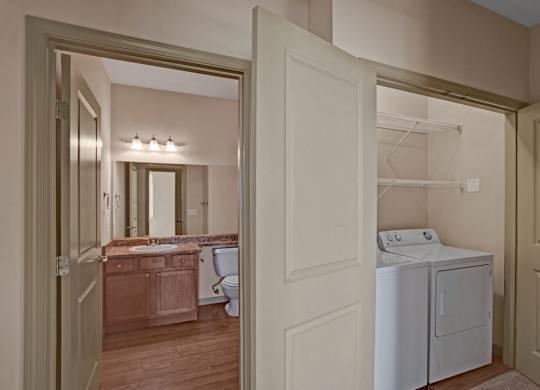 A convenient laundry area here at The Vintage with a full-size washer and dryer tucked into a closet with built-in shelving. A nearby bathroom features a wooden vanity with a speckled countertop, large mirror, and bright overhead lighting.
