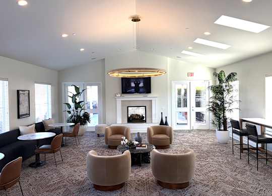 A bright resident lounge here at The Vintage with patterned carpet, cozy seating around a round coffee table, and a fireplace with a mounted TV. Large windows, tall plants, and modern lighting create a warm and welcoming gathering space.