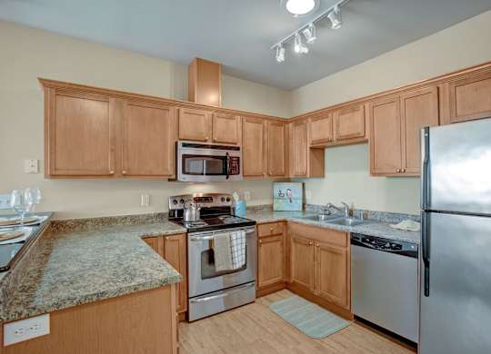 A spacious kitchen here at The Vintage with light wood cabinets, speckled granite-style countertops, and stainless steel appliances including a refrigerator, microwave, stove, and dishwasher. A double sink and bright overhead lighting create a clean, modern feel.