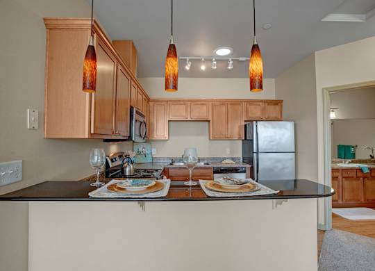 A welcoming kitchen here at The Vintage with light wood cabinets, speckled countertops, and stainless steel appliances. A raised breakfast bar with place settings sits beneath warm amber pendant lights, creating a cozy and inviting dining space.