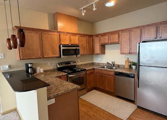 A warm kitchen here at The Vintage with rich wood cabinets, speckled countertops, and stainless steel appliances including a refrigerator, stove, microwave, and dishwasher. A raised breakfast bar with pendant lighting creates a cozy and inviting dining space.