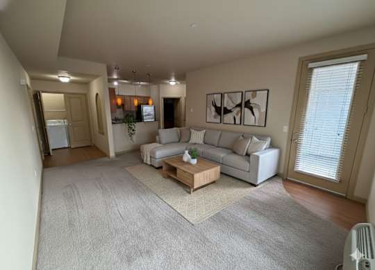 A spacious living room here at The Vintage with soft beige carpet, warm neutral walls, and a light gray sectional sofa with accent pillows. A wooden coffee table sits on a patterned rug, while a glass door with blinds leads outside.