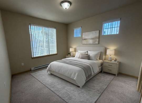 A spacious bedroom here at The Vintage with soft beige carpet, light neutral walls, and a large window with vertical blinds bringing in natural light. A bed with white bedding and neutral accent pillows sits between matching nightstands with warm lamps.