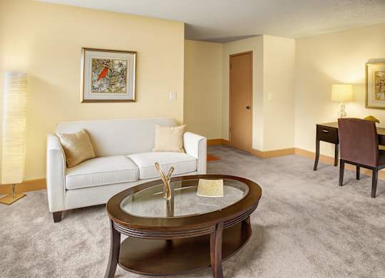 A cozy living room here at Whispering Cedars featuring a light beige sofa with neutral pillows, round glass coffee table, warm floor lamp, soft carpet, and a simple desk area creating a comfortable, inviting space.
