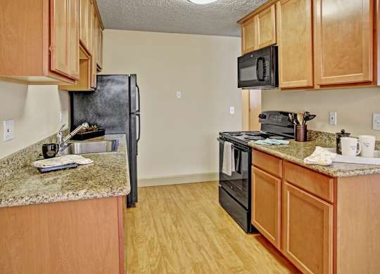 A bright kitchen here at Whispering Cedars with light brown cabinets, granite countertops, and black appliances, featuring a stainless steel sink, built-in microwave, and wood-style flooring, creating a clean, modern space with a warm, welcoming feel.