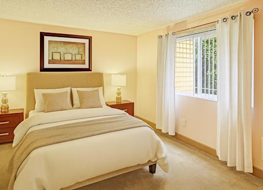 A bright bedroom here at Whispering Cedars with beige carpet, cream walls, and large windows with white curtains, featuring a comfortable bed with neutral bedding, matching bedside tables with lamps, and a warm, inviting atmosphere.