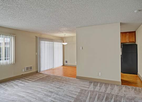 A spacious, empty living room here at Whispering Cedars with soft carpet, large windows with vertical blinds, and abundant natural light, opening to a dining area with wood-style flooring and a sliding glass door, creating a bright, open layout.