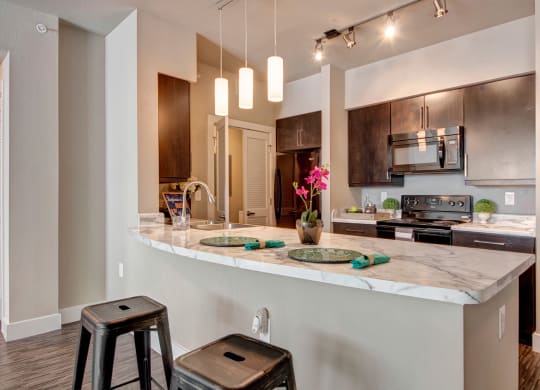 Modern kitchen here at the Enclave at Brookside Apartments with dark wood cabinets and marble countertop. Three pendant lights hang above, with two bar stools beneath. A pink orchid adds color.