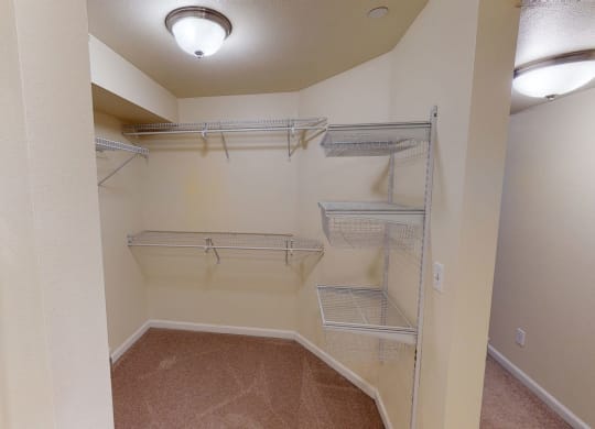 A spacious walk-in closet with beige walls and carpeted floor, featuring metal wire shelving and baskets. Soft, warm lighting from a ceiling fixture. Located at Admirals Cove Apartments.