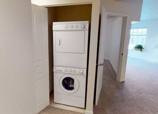 Stacked washer and dryer in a small open closet, with adjacent carpeted hallway and bright room with a large window and potted plant in the background. Located at Admirals Cove Apartments.