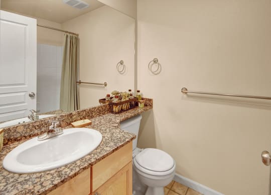 Compact bathroom with beige walls, white sink, and toilet. Granite countertop holds decorative items. Neutral shower curtain and metal towel bars. Clean and tidy ambiance. Located at Admirals Cove Apartments.