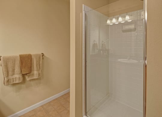 Modern bathroom with beige tiles and walls, featuring a glass shower with white tiling. Beige and brown towels hang on a towel rack, creating a cozy feel. Located at Admirals Cove Apartments.