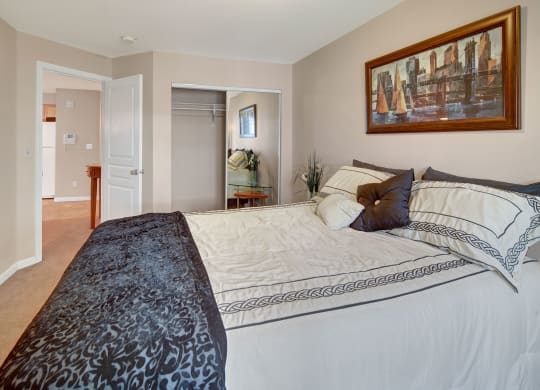 Cozy Admirals Cove Apartment bedroom with a neatly made bed featuring patterned sheets and pillows. A large painting hangs above the bed, and a closet with mirrored doors is open. Soft neutral tones create a calm ambiance.