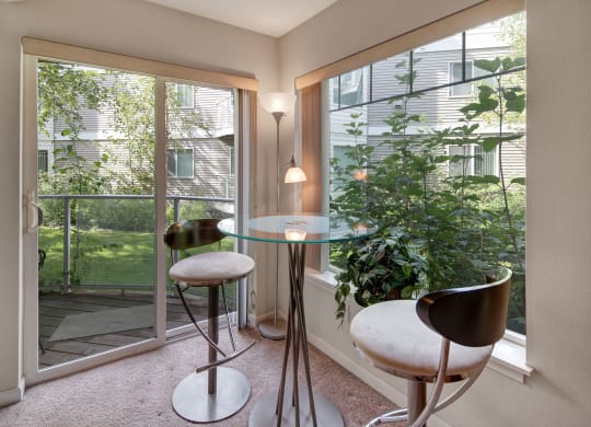 Modern dining nook with a glass table and two sleek metal stools, beside large windows overlooking lush greenery. Bright, serene atmosphere. Located at Admirals Cove Apartments.