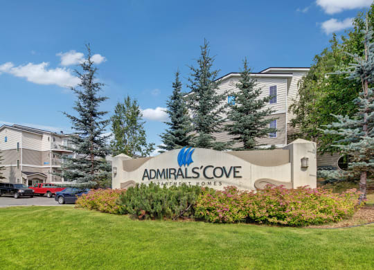 Sign for Admirals Cove Apartment Homes is surrounded by lush greenery and pine trees. Modern buildings are visible in the background under a clear blue sky.
