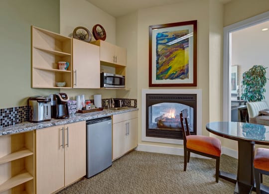 Cozy kitchen in the community lounge with light wood cabinets, granite countertop, microwave, and coffee maker. A small fridge, glass table, and lit fireplace add warmth. Located at The Alpine Apartments.