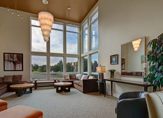 Spacious community lounge with large windows, modern chandelier, and neutral tones. Features a sectional sofa, round tables, and a decorative mirror. Airy and elegant. Located at The Alpine Apartments.