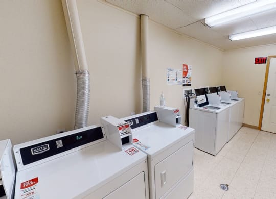 Small laundry room with six white washing machines and dryers, beige walls, a door with a window, fluorescent lights, and a red exit sign. Amber Ridge Apartments.