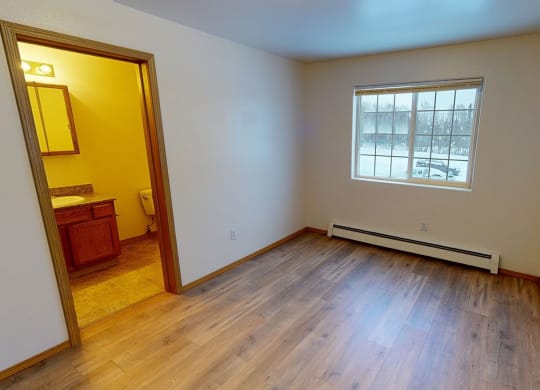 Empty bedroom with light wood flooring, cream walls, and a large window showing snowy trees. Doorway leads to a brightly lit bathroom. Amber Ridge Apartments.